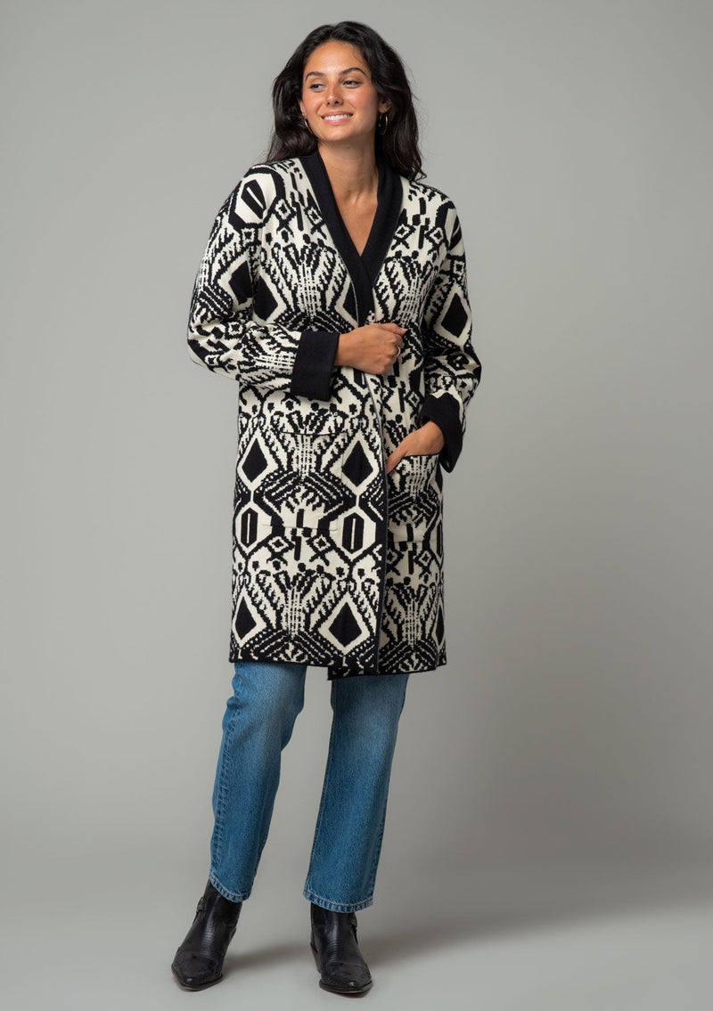 [Color: Natural/Black] A full body front facing image of a brunette model wearing a natural off white and black bohemian patterned mid length cardigan. A long sweater style with long sleeves, contrast black wrist cuffs, a contrast black shawl collar, an open front, and side pockets. 