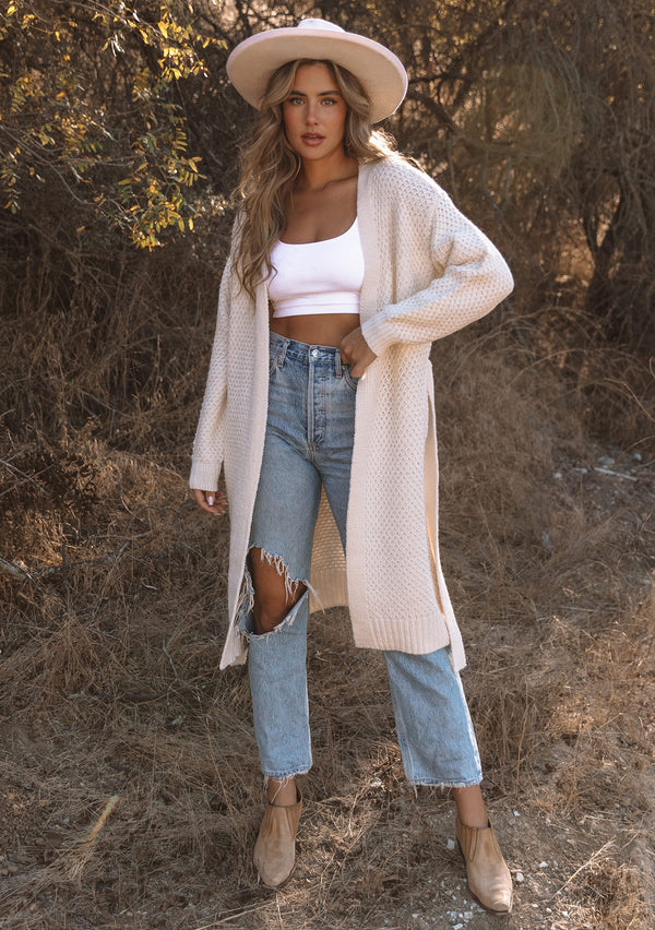 [Color: Natural] A full body front facing image of a blonde model wearing a natural mid length cardigan in a textured honeycomb knit. With long sleeves, a tie front waist belt, and side pockets.
