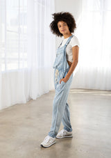 [Color: Reagan Wash] A model wearing classic light wash overalls. With a drawstring waist, side pockets, adjustable straps, a full length leg, and a zippered front pocket.