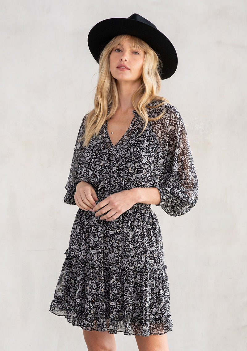 [Color: Black/Natural] A model wearing a cute bohemian paisley chiffon mini dress featuring a smocked elastic waist and a split v neckline with tassel ties. With gold lurex detail.