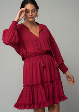 [Color: Burgundy] A front facing image of a burgundy red bohemian mini dress in a textured shadow stripe. With voluminous long dolman sleeves and smocked elastic wrist cuffs, a split v neckline with tassel ties, a ruffle trimmed tiered skirt, and a smocked elastic waist. 