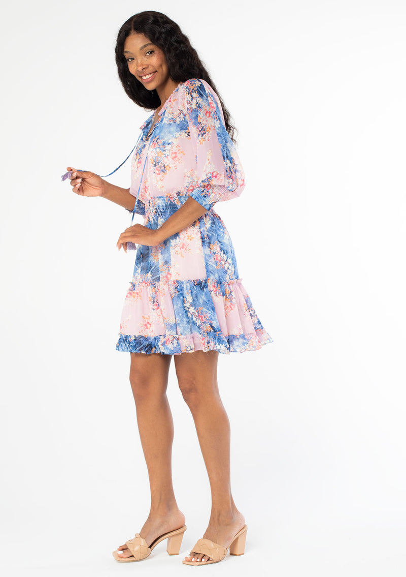[Color: Lavender/Denim Blue] A full body side facing image of a black model wearing a bohemian lavender purple and blue floral print chiffon mini dress with long sleeves and tassel neck ties. 