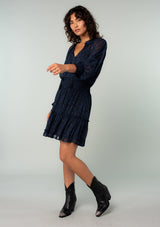 [Color: Black/Blue] A side facing image of a brunette model wearing a black and navy blue paisley print chiffon mini dress with silver clip dot thread accents throughout. With long sheer sleeves, a smocked elastic waist, a split ruffled neckline with tassel ties, and a flowy ruffle trimmed tiered mini skirt.