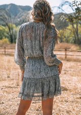[Color: Grey/Natural] A back facing image of a blonde model wearing a best selling bohemian mini dress in sheer chiffon, designed in a grey and natural paisley print. With long sleeves, smocked elastic wrist cuffs, a smocked elastic waist, ruffle trimmed tiered skirt, and tassel tie neckline. 