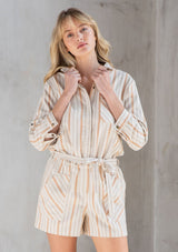 [Color: Natural/Terracotta] A model wearing a linen blend twill short romper in a terracotta yarn dye stripe. With long rolled tab sleeves and a tie waist belt. 
