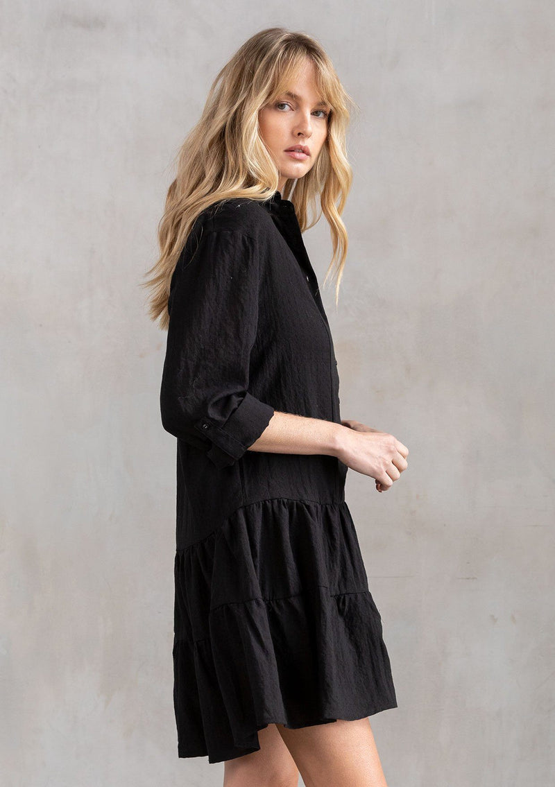[Color: Black] A model wearing a black baby doll mini dress with a button front, a classic collared neckline, long rolled sleeves with a button tab closure, and a tiered skirt.