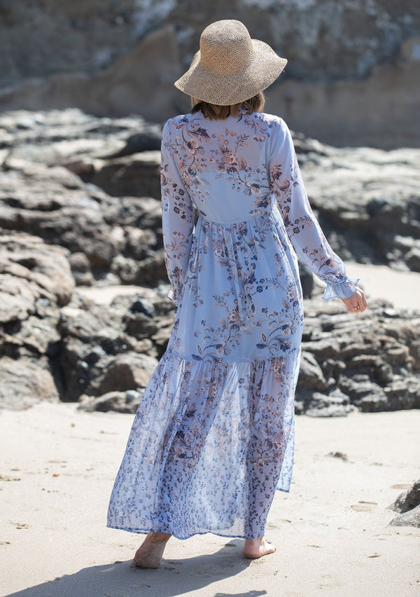 [Color: Light Blue] A woman on the beach wearing a dreamy vintage floral maxi dress. Featuring long voluminous sheer sleeves with a flounce wrist cuff, a button front, and an empire waist.