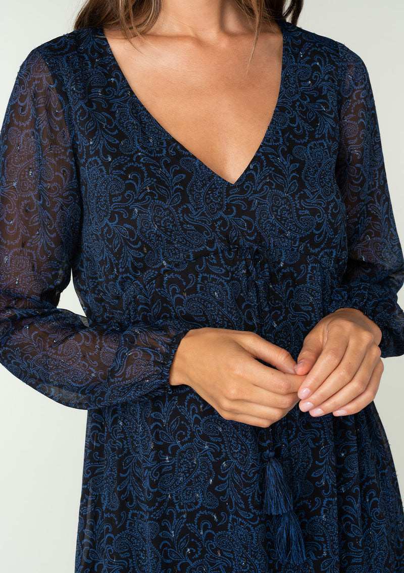 [Color: Black/Blue] A close up front facing image of a brunette model wearing a best selling navy blue chiffon maxi dress with black paisley print and light catching metallic clip dot details. Perfect for special occasions, with sheer long sleeves, a v neckline, an empire waist with drawstring tassel tie detail, and an asymmetric ruffled trimmed tiered skirt. 