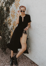 [Color: Black] A woman standing outside wearing a black mid length shirt dress in a linen blend. Featuring short sleeves with a ruffled trim, a button up front, an adjustable waist tie, and a ruffled hemline.