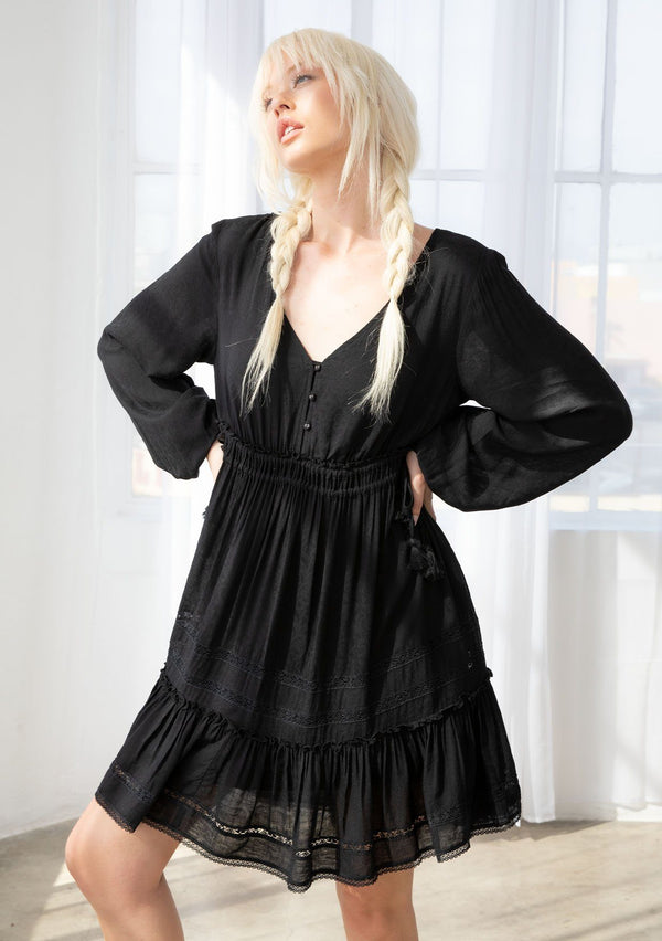 [Color: Black] A blond model wearing a black bohemian mini dress in a textured paisley jacquard. With a loop button front top, ruffle trimmed waist with adjustable tassel tie drawstring, a lace trimmed skirt, and long volume sleeves.  