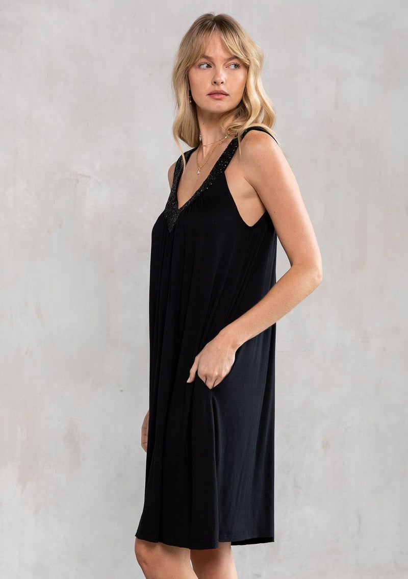[Color: Black] A model wearing a black soft knit, holiday party mini dress with beaded straps, a deep v neckline in front and back, and a flowy swing silhouette. 