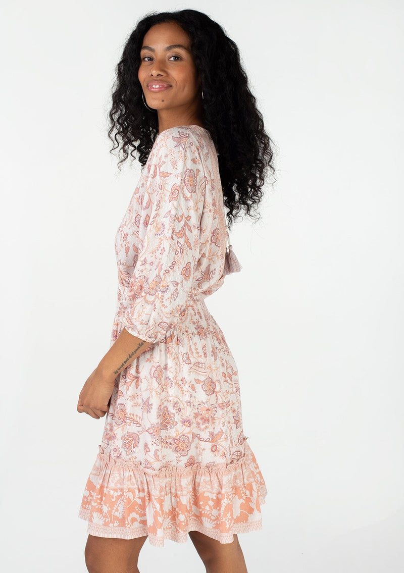 [Color: Natural/Clay] A side facing image of a brunette model wearing a bohemian spring mini dress in a pink floral border print. With voluminous three quarter length sleeves, a v neckline, a smocked elastic waist, a ruffle trimmed flowy tiered skirt, and an open back with tassel tie closure. 