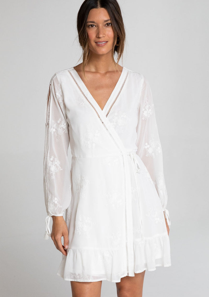 [Color: White] A model wearing a charming white embroidered chiffon mini wrap dress, with sheer long split sleeves, lattice trim details, and a wrap front with side tie closure.