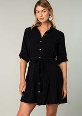 [Color: Black] A front facing image of a blonde model wearing a black cotton jacquard mini shirt dress. An elevated bohemian short dress with a swingy tiered skirt, a button front, a classic collared neckline, a self tie waist belt, and long rolled sleeves with a button tab closure. 