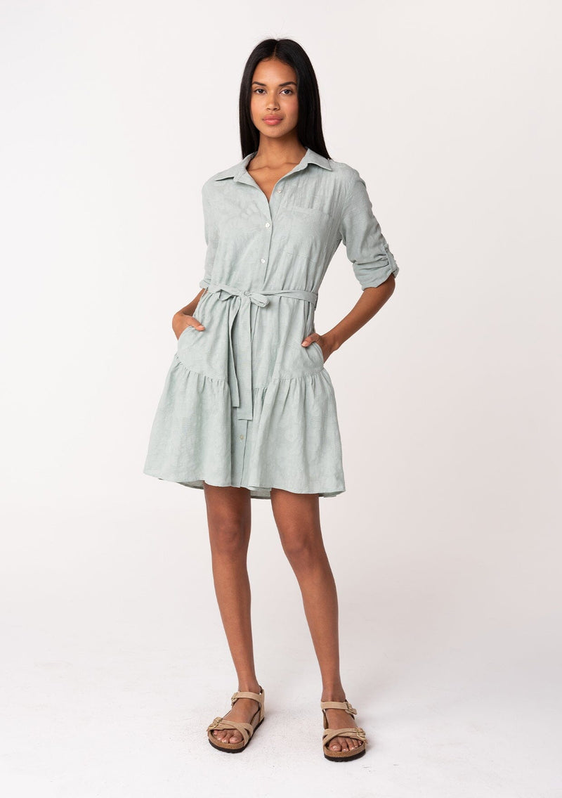 [Color: Sage] A full body front facing image of a brunette model wearing a light sage green cotton jacquard mini shirt dress. An elevated bohemian short dress with a swingy tiered skirt, a button front, a classic collared neckline, a self tie waist belt, and long rolled sleeves with a button tab closure.