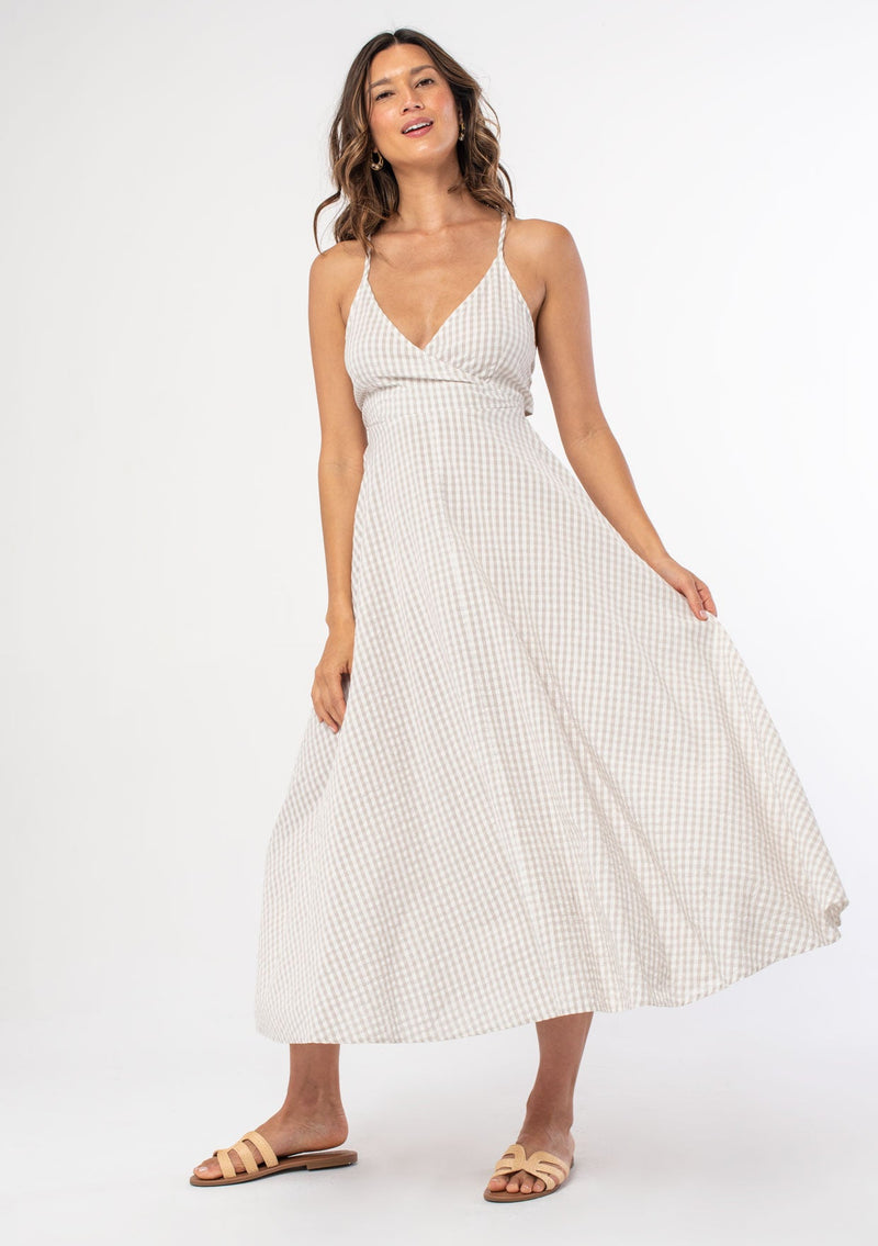 [Color: Pebble/Ivory] A woman wearing a cotton tan and off white gingham check mid length dress with adjustable spaghetti straps and a tie back detail. 