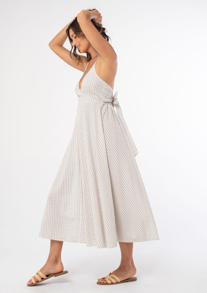 [Color: Pebble/Ivory] A woman wearing a cotton tan and off white gingham check mid length dress with adjustable spaghetti straps and a tie back detail. 
