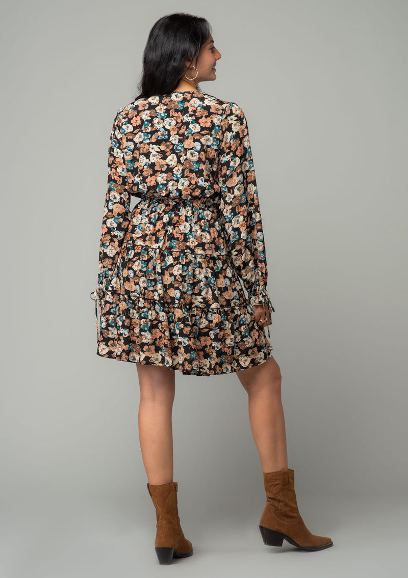 [Color: Black/Natural] A back facing image of a brunette model wearing a bohemian mini dress in a black and natural floral print. With long sleeves, adjustable wrist cuffs, and a flowy tiered mini skirt. 