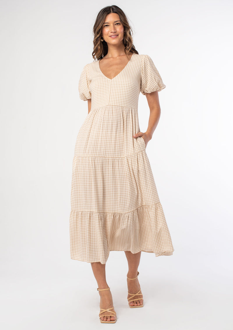 [Color: Tan/Natural] A model wearing a tan and white small checkered gingham mid length dress with short puff sleeves and an open back detail with button closure.