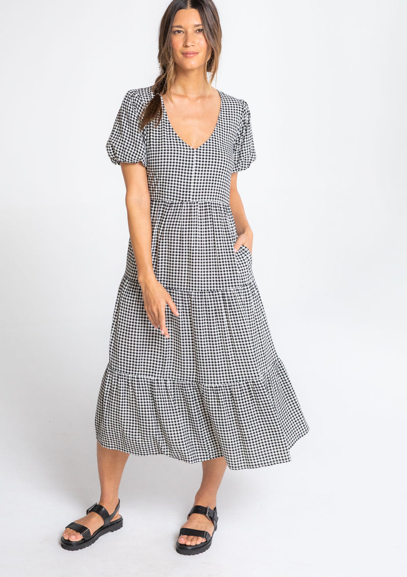 [Color: Black/Natural] A model wearing a black and white small checkered gingham mid length dress with short puff sleeves and an open back detail with button closure. 