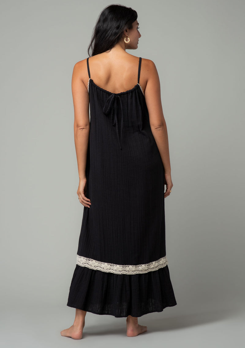 [Color: Black/Natural] A full body back facing image of a brunette model wearing a black cotton gauze sleeveless maxi dress with a natural crochet trim top and hemline. With adjustable spaghetti straps and a flowy fit. 