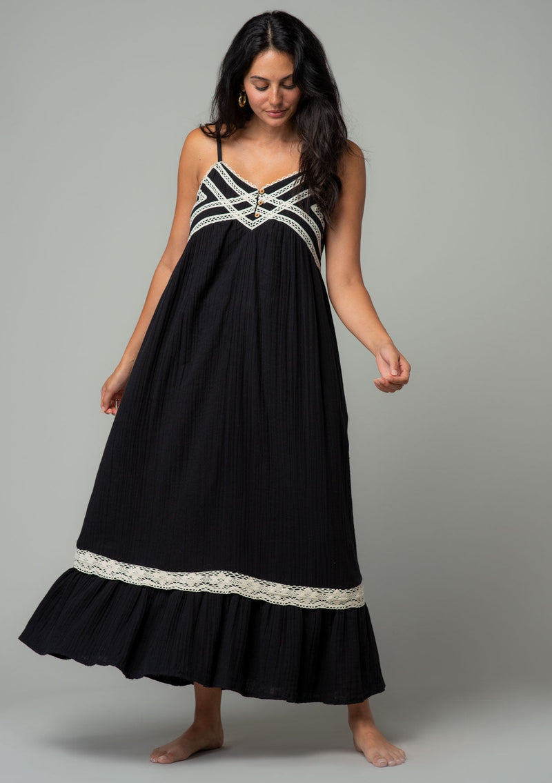 [Color: Black/Natural] A full body front facing image of a brunette model wearing a black cotton gauze sleeveless maxi dress with a natural crochet trim top and hemline. With adjustable spaghetti straps and a flowy fit. 