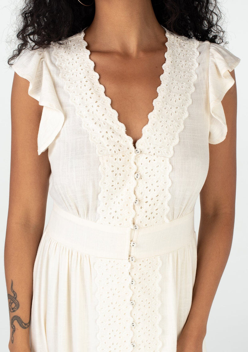 [Color: Vanilla] A close up front facing image of a brunette model wearing a classic ivory bohemian spring maxi dress with eyelet lace trim throughout. Featuring short flutter cap sleeves, a v neckline, a self covered loop button front, and a tiered flowy skirt. 
