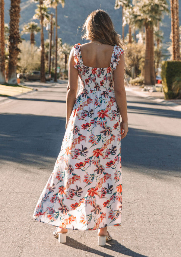 [Color: Ivory/Red] A model wearing a white bohemian maxi dress with pink and red tropical floral print. With a slim fit smocked bodice and a flowy skirt. 