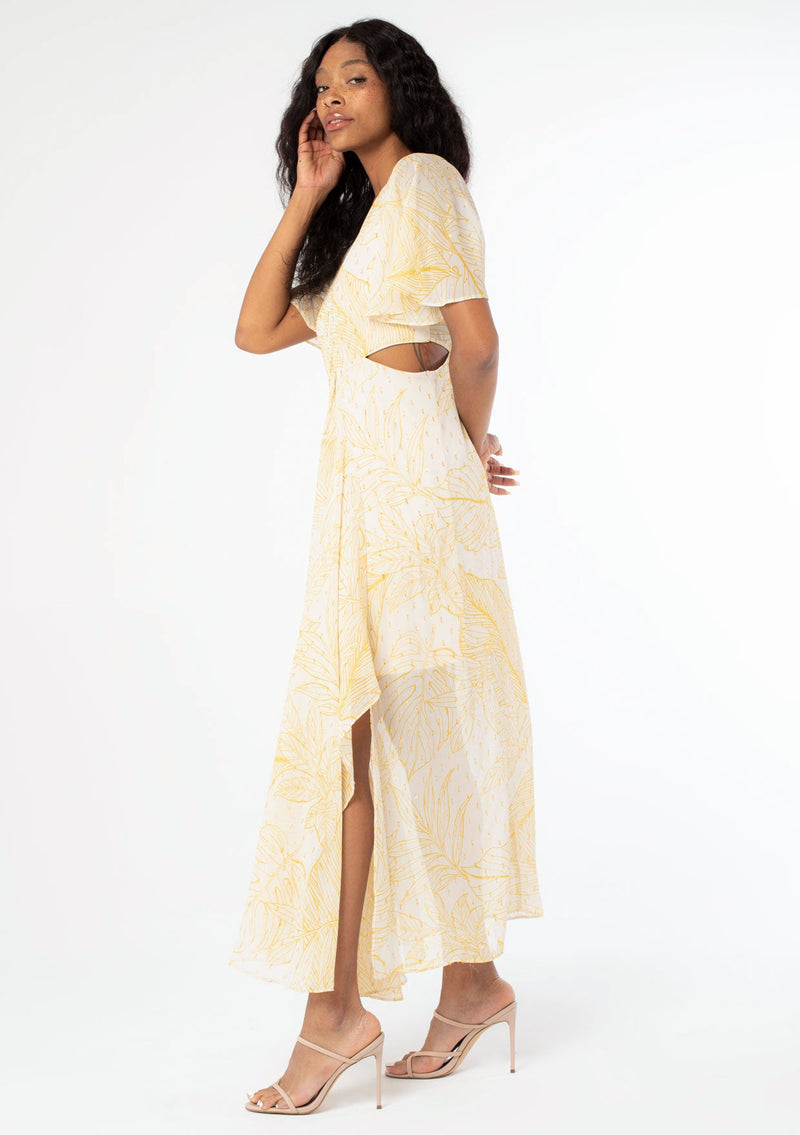[Color: Natural/Mustard] A model wearing a yellow leaf print maxi dress in sheer chiffon. With metallic gold clip dot details, side waist cutouts, and short flutter sleeves.