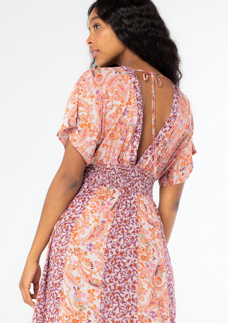 [Color: Lavender/Orange] A close up back image of a black model with long dark wavy hair wearing a purple and orange mixed floral print maxi dress with short kimono sleeves, a smocked elastic waist, and a side slit. 