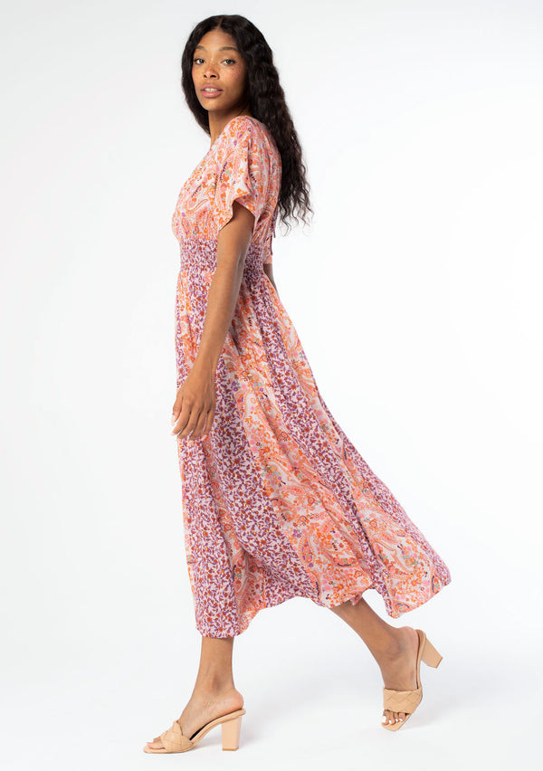 [Color: Lavender/Orange] A walking side image of a black model with long dark wavy hair wearing a purple and orange mixed floral print maxi dress with short kimono sleeves, a smocked elastic waist, and a side slit. 