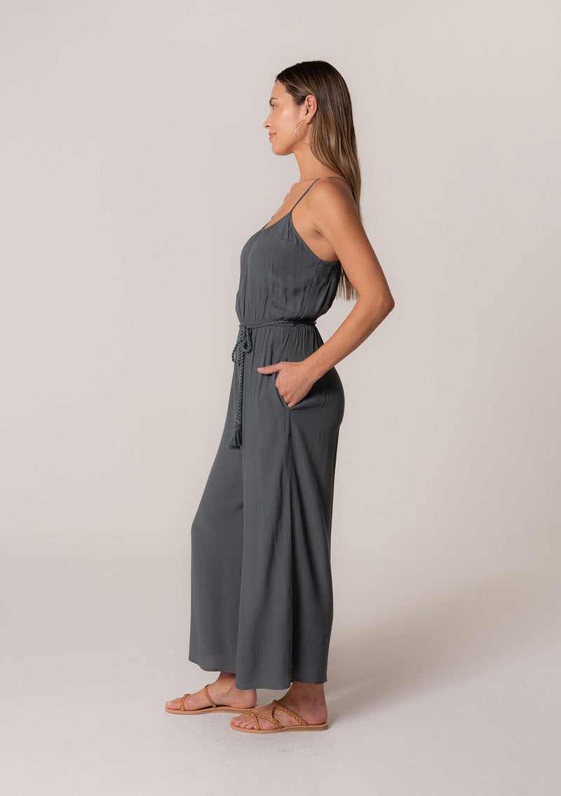 [Color: Slate] A side facing image of a brunette model wearing a solid blue bohemian one piece jumpsuit. With adjustable spaghetti straps, a scoop neckline, an elastic waist, an adjustable braided rope belt with tassel accents, and a long wide leg.
