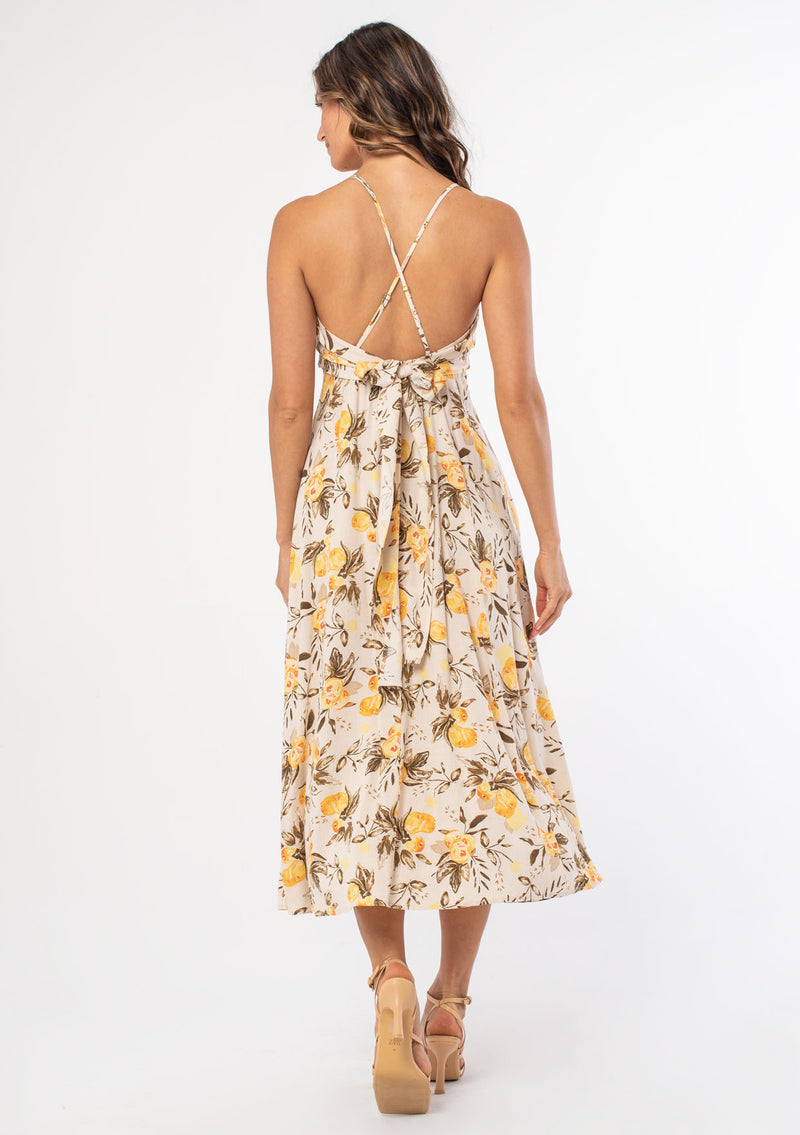 [Color: Natural/Yellow] A model wearing a natural and yellow fruit floral print mid length dress with spaghetti straps and a tie back detail. 