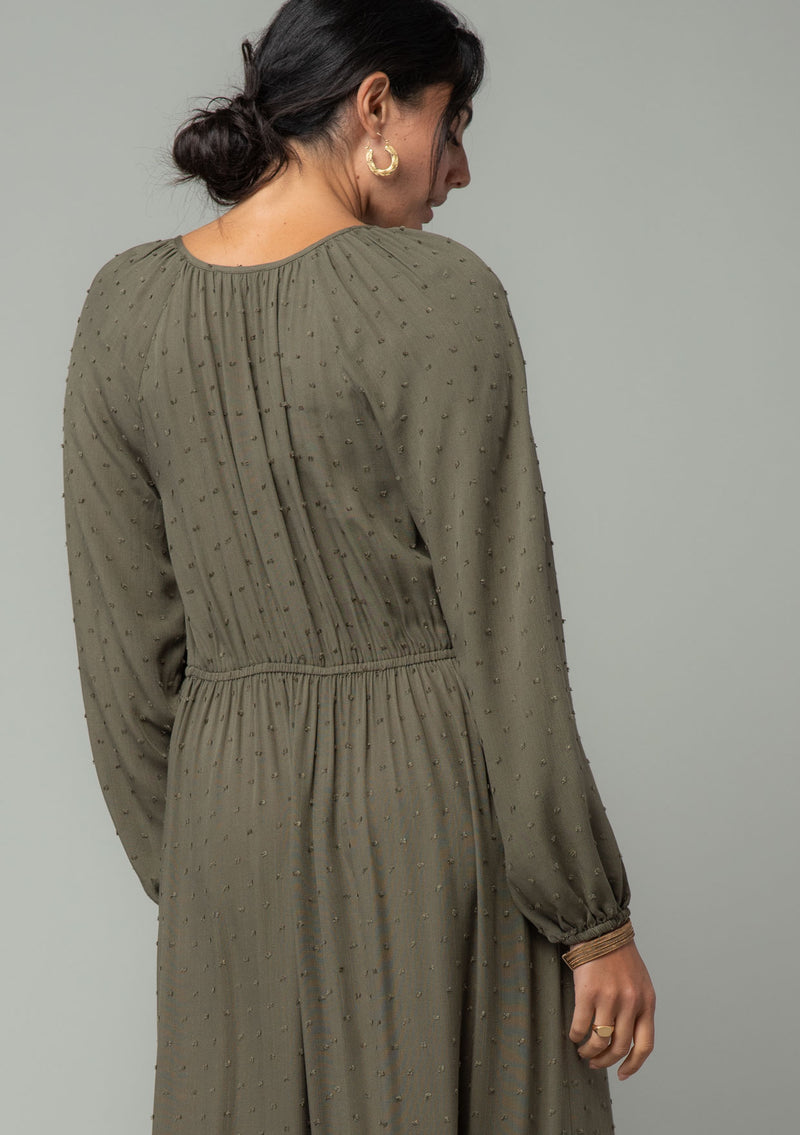[Color: Olive] A half body back facing image of a brunette model wearing an olive green high low maxi dress with a ruffled hemline, voluminous long sleeves, and a drawstring tassel tie waist. 