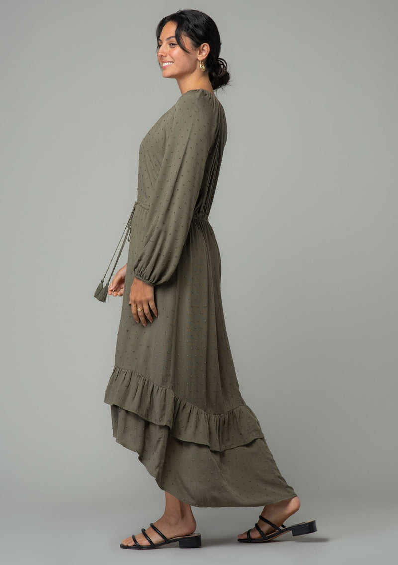 [Color: Olive] A side facing image of a brunette model wearing an olive green high low maxi dress with a ruffled hemline, voluminous long sleeves, and a drawstring tassel tie waist. 