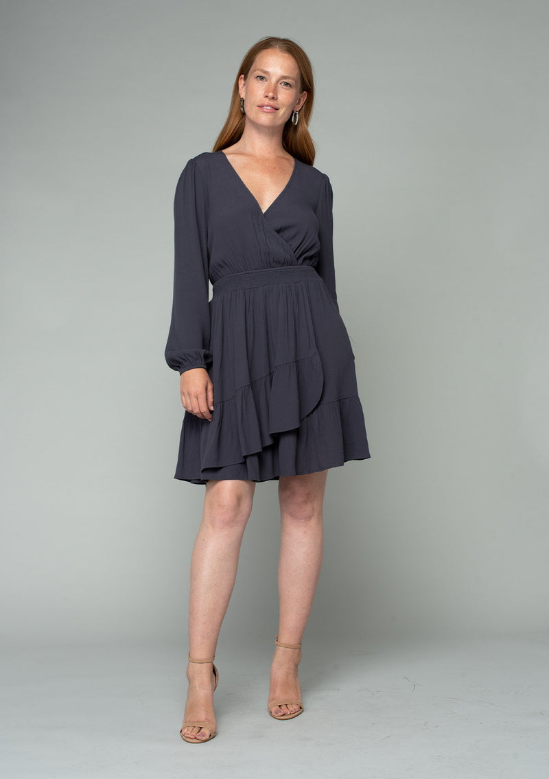 [Color: Charcoal] A full body front facing image of a red headed model wearing a charcoal grey bohemian mini dress designed in a lightweight crepe. With long sleeves, a surplice v neckline, and a ruffled faux wrap skirt. 