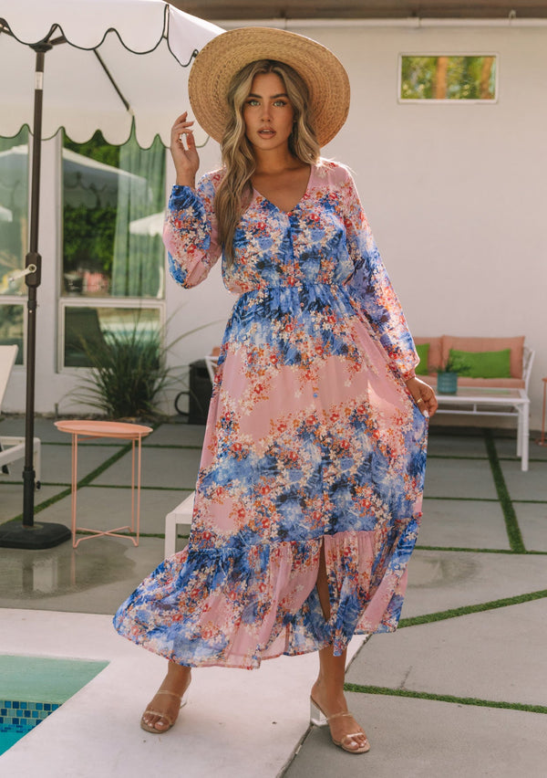 [Color: Lavender/Denim Blue] A moving front facing image of a blonde model wearing a lavender purple and blue floral print bohemian chiffon maxi dress. With long sleeves, a self covered button front, and an elastic waist.