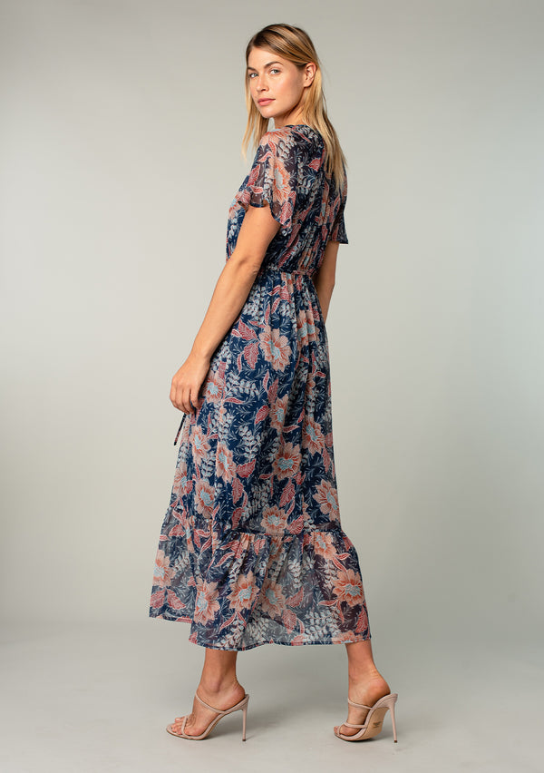 [Color: Navy/Rust] A side facing image of a blonde model wearing a sheer chiffon bohemian maxi dress in a navy blue and rust red floral print. Wit short flutter sleeves, a long tiered skirt, and an adjustable drawstring waist.