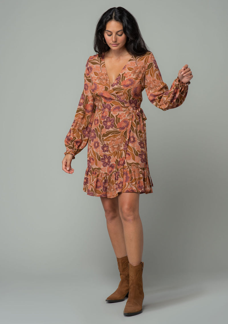 [Color: Clay/Olive] A full body front facing image of a brunette model wearing a clay brown and olive green retro floral print mini wrap dress. With long sleeves, a ruffled hemline, and a side tie closure. 