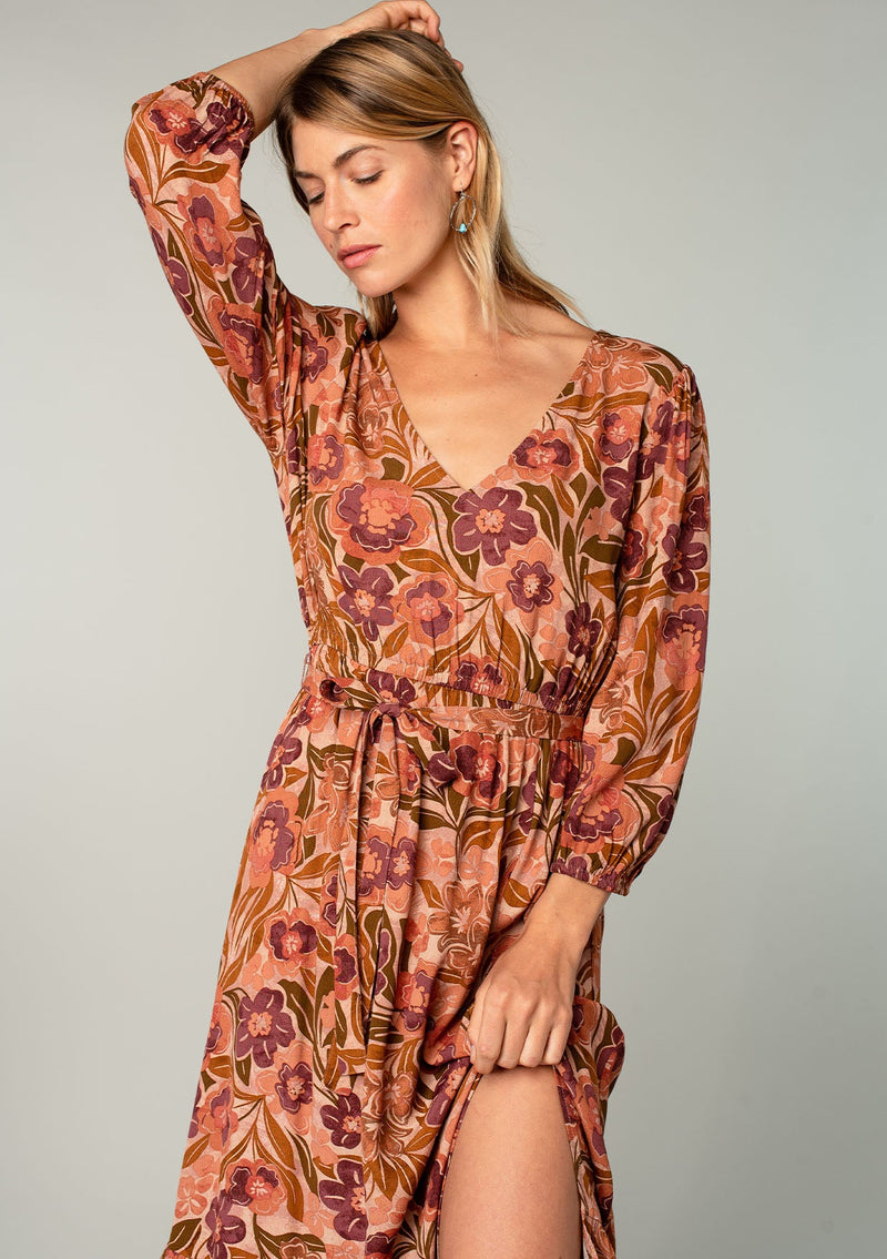 [Color: Clay/Olive] A close up front facing image of a blonde model wearing a clay brown and olive green retro floral print maxi dress. With long sleeves, a side slit, and a tie waist. 