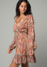 [Color: Natural/Rust] A side facing image of a brunette model wearing a bohemian mini dress in a natural and rust red paisley floral print. With long sleeves, a smocked elastic waist, and a back keyhole with tie closure. 