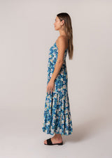 [Color: Dusty Teal/Blue] A side facing image of a brunette model wearing a flowy summer maxi dress in a blue floral print. With adjustable spaghetti straps, a scalloped edge v neckline, a button front top, a flowy tiered skirt, and side pockets. 