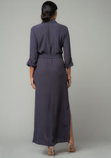 [Color: Charcoal] A back facing image of a brunette model wearing a relaxed fit charcoal grey maxi shirt dress made from a lightweight crepe. With long rolled sleeves, a button tab sleeve closure, two front pockets, side pockets at the hip, and a self tie waist belt. 