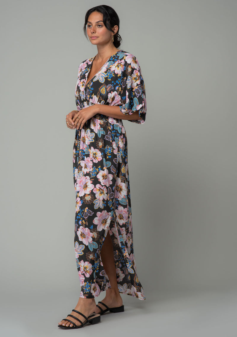 [Color: Charcoal/Rose] A side facing image of a brunette model wearing a charcoal grey and pink floral print bohemian resort maxi dress with half length kimono sleeves, an open back with tie closure, and a smocked elastic waist. 