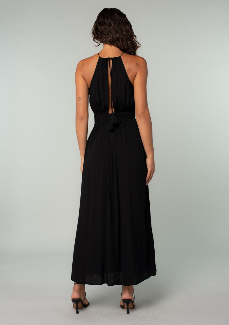 [Color: Black] A back facing image of a brunette model wearing a bohemian black halter maxi dress. A holiday maxi dress with thin spaghetti straps, a back keyhole, a halter neckline with adjustable back tassel ties, light catching beaded accents, a half smocked elastic waist at the back, and a long flowy skirt. 