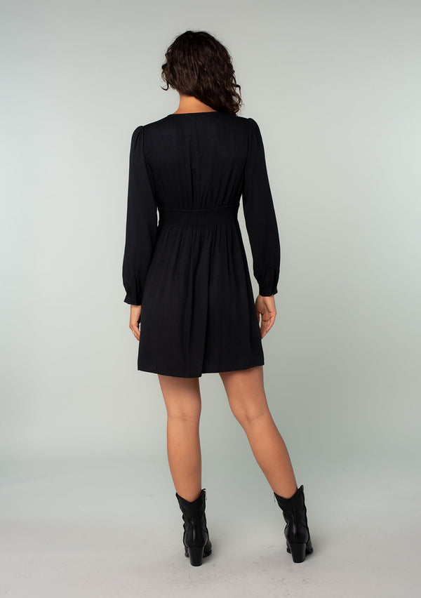 [Color: Black] A back facing image of a brunette model wearing a black long sleeve mini dress. Perfect for the holidays, with a self covered button up front, a v neckline, and a half smocked elastic waist at the back.