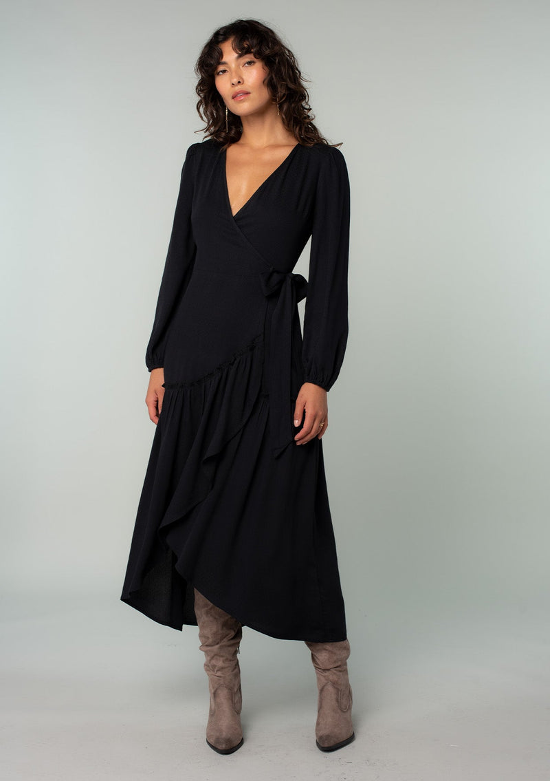[Color: Black] A front facing image of a brunette model wearing a black maxi wrap dress with long sleeves, a ruffle trimmed tiered high low hemline, and a side tie closure. 