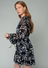 [Color: Black/Lavender] A side facing image of a red headed model wearing a sheer chiffon bohemian mini dress in a black and lavender purple floral print. With a ruffle trimmed tiered skirt, an elastic waist, a v neckline, and long flared sleeves with adjustable drawstring wrist ties. 