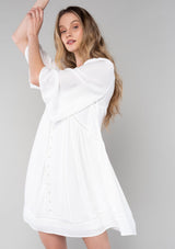 [Color: Vanilla] A half body front facing image of a blonde model wearing a bohemian white flowy baby doll mini dress. A spring dress with half length flared sleeves, a v neckline, a self covered button front, and lattice trim. 