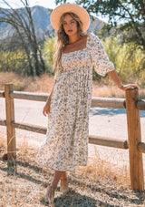 [Color: Natural/Black] A front facing image of a blonde model wearing a bohemian mid length dress in an off white and black ditsy floral print. With three quarter length sleeves, wrist ties, a slim fit smocked bodice, a square neckline, and an empire waist.
